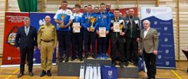 „Na zdjęciu: drużyny strażaków – zwycięzcy klasyfikacji drużynowej 41. Mistrzostw Polski Strażaków PSP w tenisie stołowym w Suchedniowie – stoją na podium z pucharami i dyplomami.