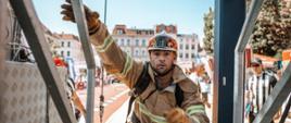 Strażak w umundurowaniu specjalnym wchodzi po schodach – widać jego popiersie, prawą ręką chwyta poręcz. W tle w tle zabudowa kamieniczna miasta.