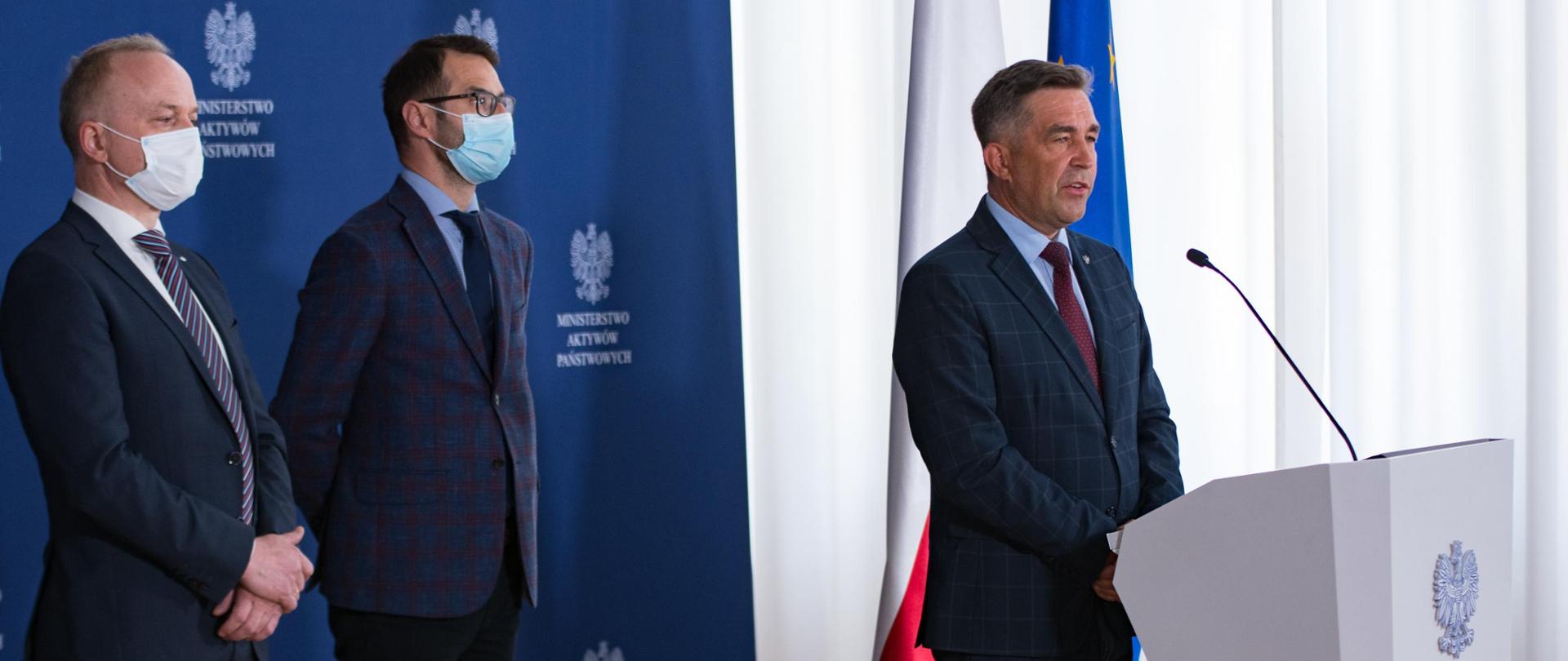 Boczne ujęcie: wiceminister Zbigniew Gryglas przy mównicy. Za nim stoją Czesław Warsewicz i Tomasz Poręba. W tle ścianka z logotypami MAP oraz flagi polska i unijna. 
