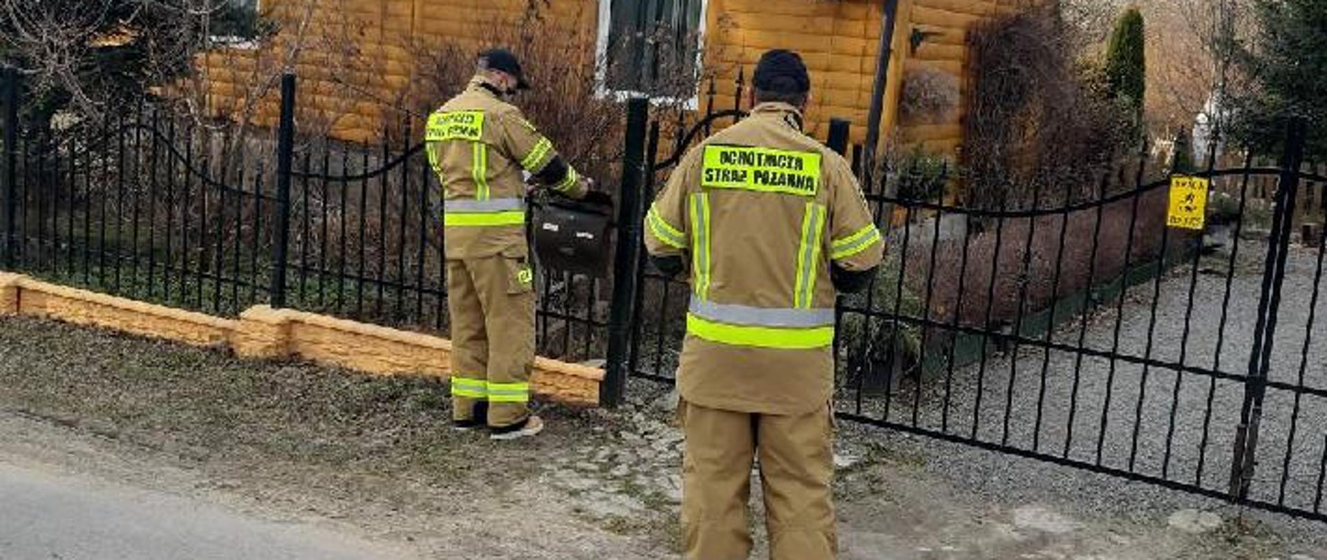 Zdjęcie przedstawia 2 strażaków OSP stojących przed bramą do posesji. Strażacy wkładają ulotkę informacyjną do skrzynki na listy