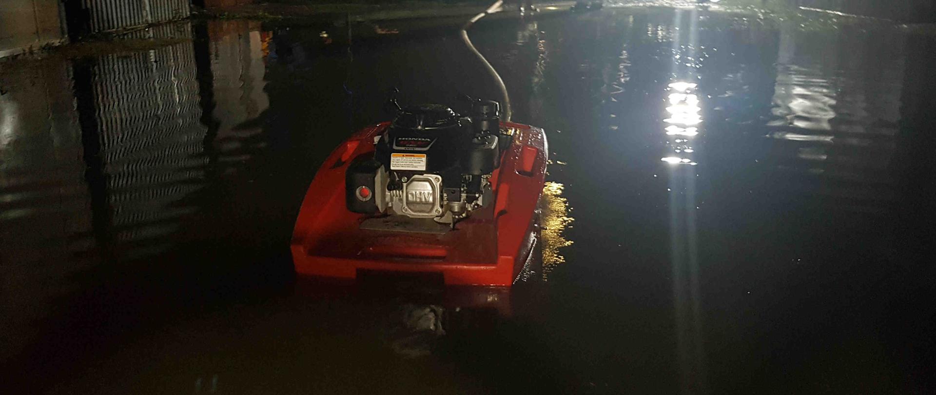 Zdjęcie zrobione w nocy. Na zdjęciu widać rozlewisko wodne. Strażacy pompują wodę przy pomocy pompy. 