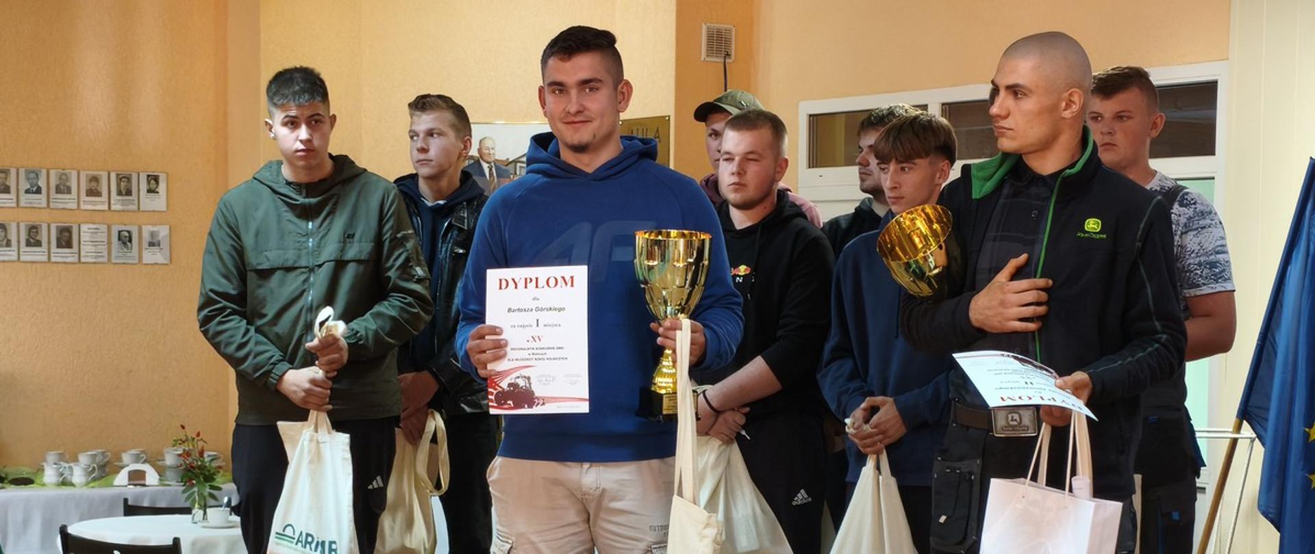 Uczeń w niebieskiej bluzie i jasnych spodniach stoi z pucharem i dyplomem. W tle 8 chłopców. Przed wszystkimi na ziemi stoją nagrody rzeczowe.