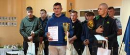Uczeń w niebieskiej bluzie i jasnych spodniach stoi z pucharem i dyplomem. W tle 8 chłopców. Przed wszystkimi na ziemi stoją nagrody rzeczowe.