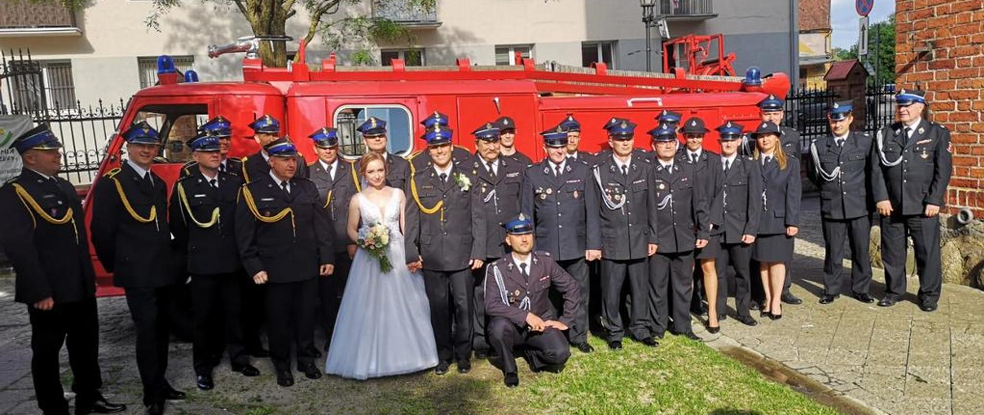 Strażacy w mundurach galowych z para młodą na tle zabytkowego samochodu strażackiego.