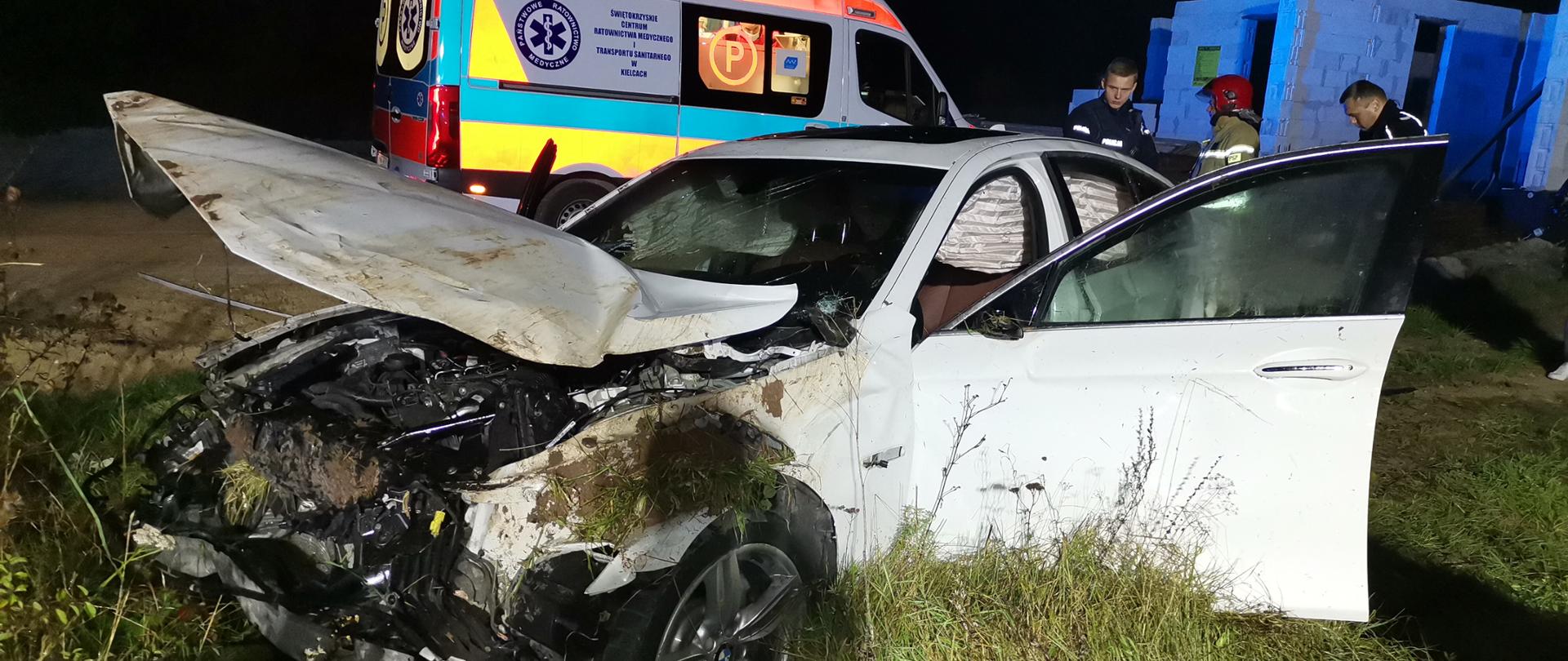 Zdjęcie przedstawia zniszczony samochód osobowy po dachowaniu. Za autem stoi ambulans Państwowego Ratownictwa Medycznego, a z prawej strony widać dwóch funkcjonariuszy Policji oraz strażaka.
