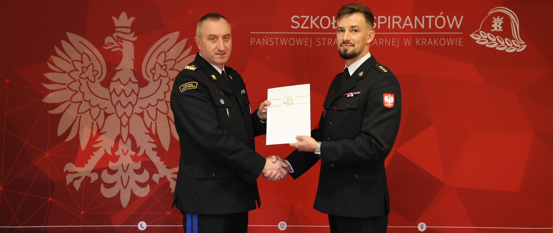 Komendant nadbryg. Piotr Filipek po lewej stronie ściska dłoń uśmiechniętego st. kpt. Wojciecha Brody po prawej, wręczając mu jednocześnie białą teczkę z aktem mianowania. Tło stanowi czerwona ścianka promocyjna.