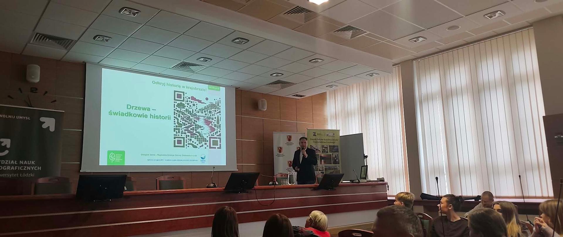 Prelegent prowadzi prezentację pt. „Drzewa – świadkowie historii” podczas spotkania z okazji Dnia Krajobrazu. Na ekranie wyświetlony jest slajd prezentacji, a na pierwszym planie widoczni są uczestnicy wydarzenia siedzący w sali konferencyjnej.