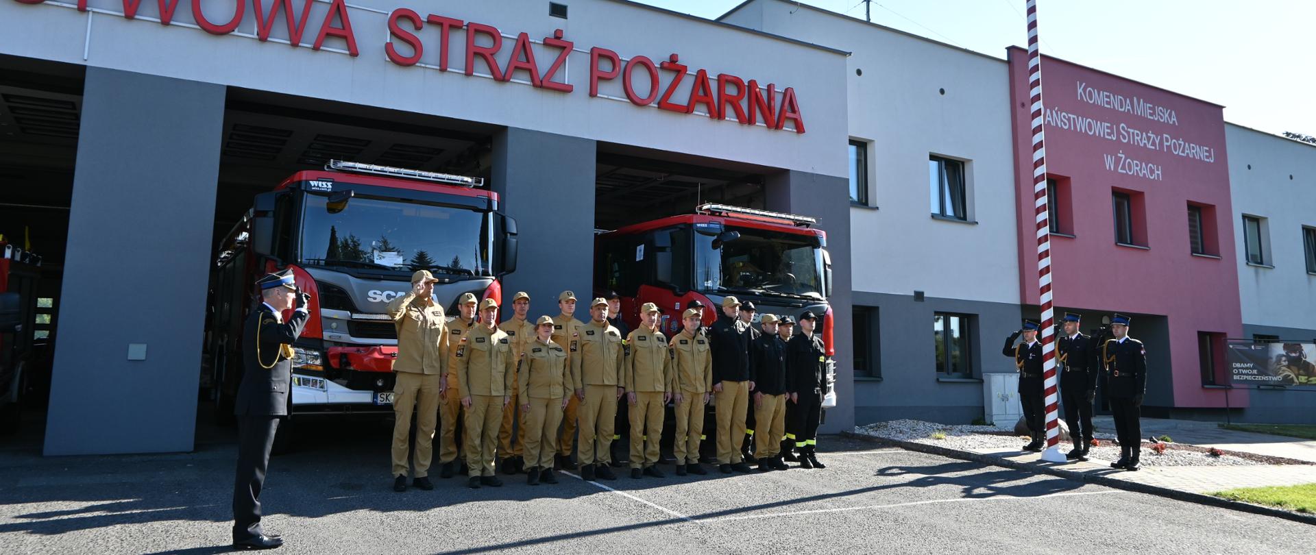 Grupa strażaków stojących przed budynkiem straży pożarnej. Na budynku widnieje napis "PAŃSTWOWA STRAŻ POŻARNA". Przed strażakami stoi jeden strażak w mundurze, który salutuje. W tle widać dwa czerwone wozy strażackie. Po prawej stronie zdjęcia znajduje się maszt z flagą Polski oraz kilku strażaków salutujących. Na budynku po prawej stronie widnieje napis "KOMENDA MIEJSKA PAŃSTWOWEJ STRAŻY POŻARNEJ W ŻORACH".