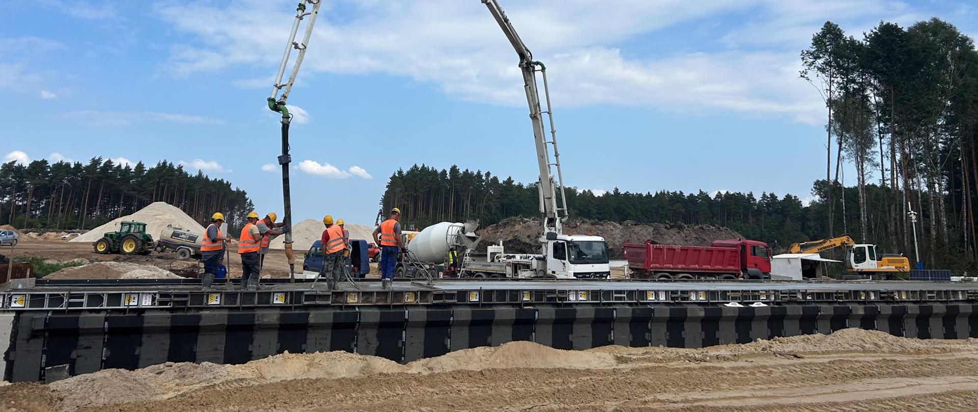Budowa S74 Mniów - Kielce - Betonowanie przepustu, robotnicy na konstrukcji obiektu trzymają wysięgnik pompy do betonu, za obiektem samochody ciężarowe i betoniarka 