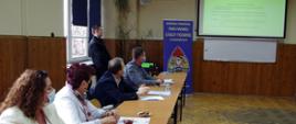 Widok z boku. Na sali szkoleniowej na pierwszym planie przy stolikach siedzą uczestnicy spotkania. Na drugim planie stoi strażak PSP referujący zagadnienia. W tle widoczny ekran oraz roll-up z logo KP PSP w Wadowicach.