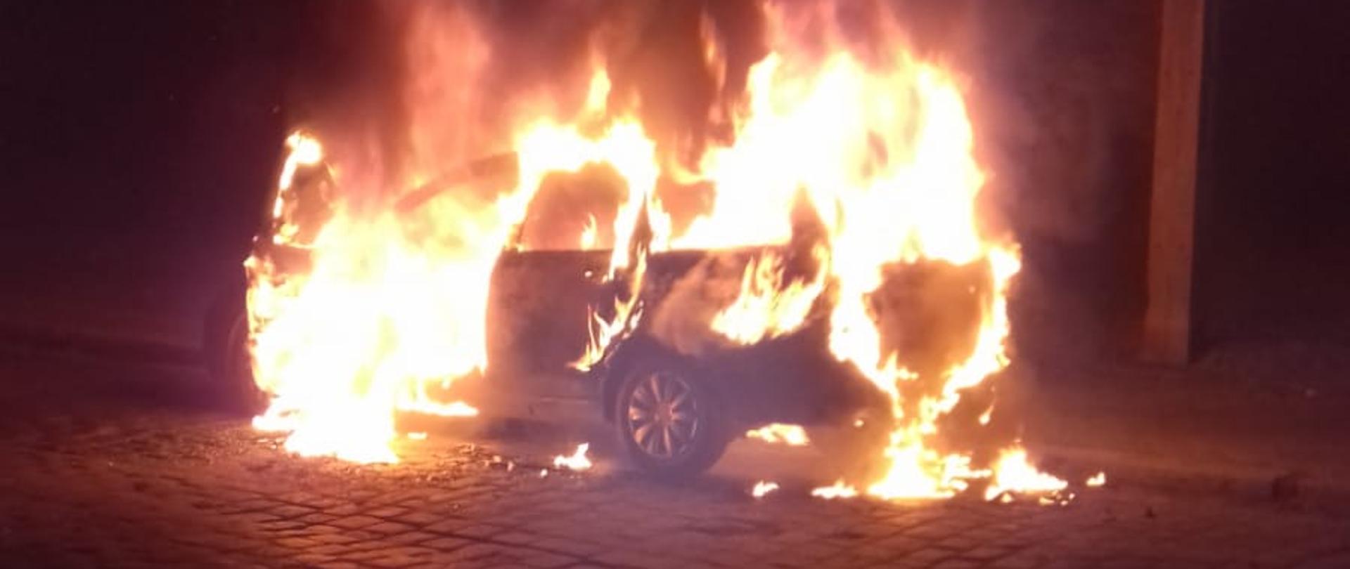 Zdjęcie przedstawia samochód osobowy z otwartymi przednimi drzwiami kierowcy, który w całości objęty jest pożarem. Pojazd znajduje się na brukowej drodze dojazdowej