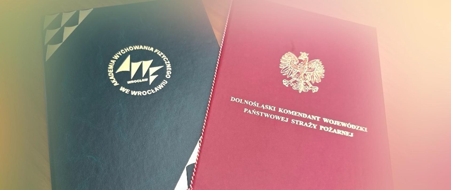 Dwaj mężczyźni podpisują oficjalne dokumenty, a następnie pozują z teczkami oraz podają sobie dłonie na tle zielonego baneru z logo Akademii Wychowania Fizycznego we Wrocławiu; jeden z nich jest ubrany w galowy munduru Państwowej Straży Pożarzanej, a drugi w elegancki garnitur.