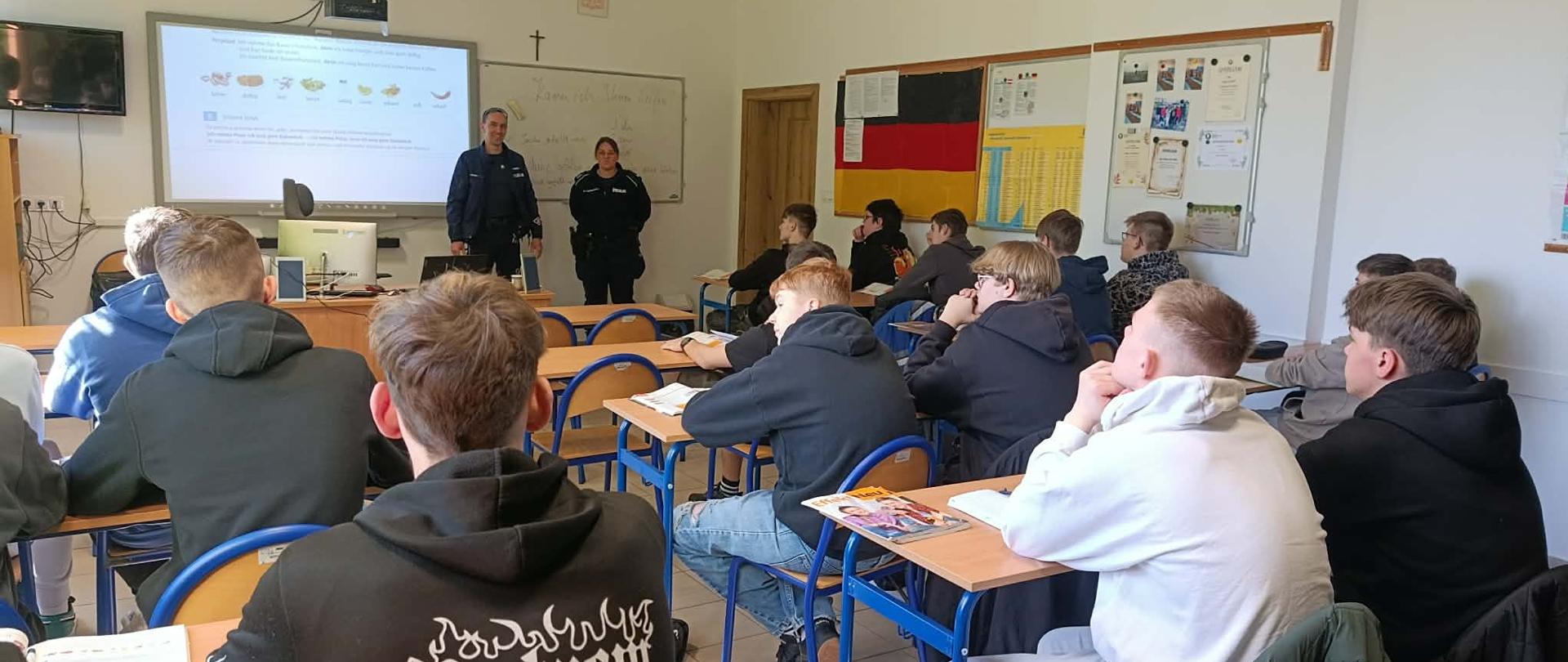 Uczniowie siedzą w klasie, przy tablicy stoją policjanci - kobieta i mężczyzna, którzy prowadzą wykład