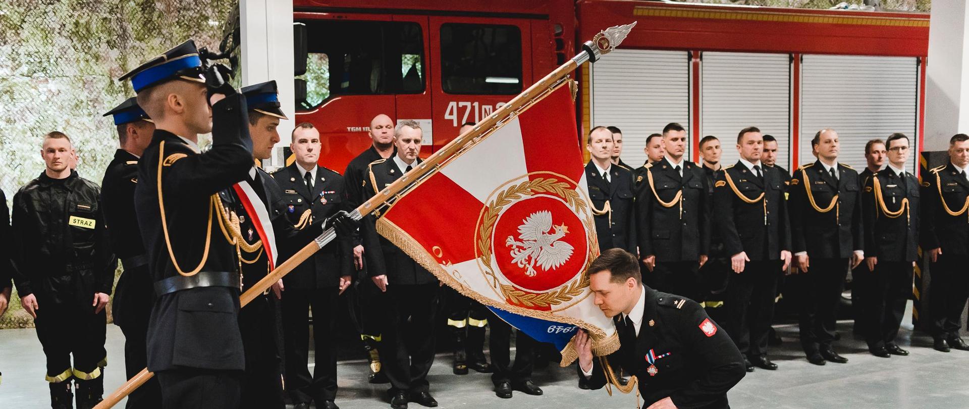 Zdjęcie przedstawia Komendanta Powiatowego PSP w Kętrzynie mł. bryg. Kamila Golon klęczącego na jednym kolanie przed sztandarem PSP Kętrzyn. Za nim znajdują się dwa czerwone pojazdy KP PSP Kętrzyn. 