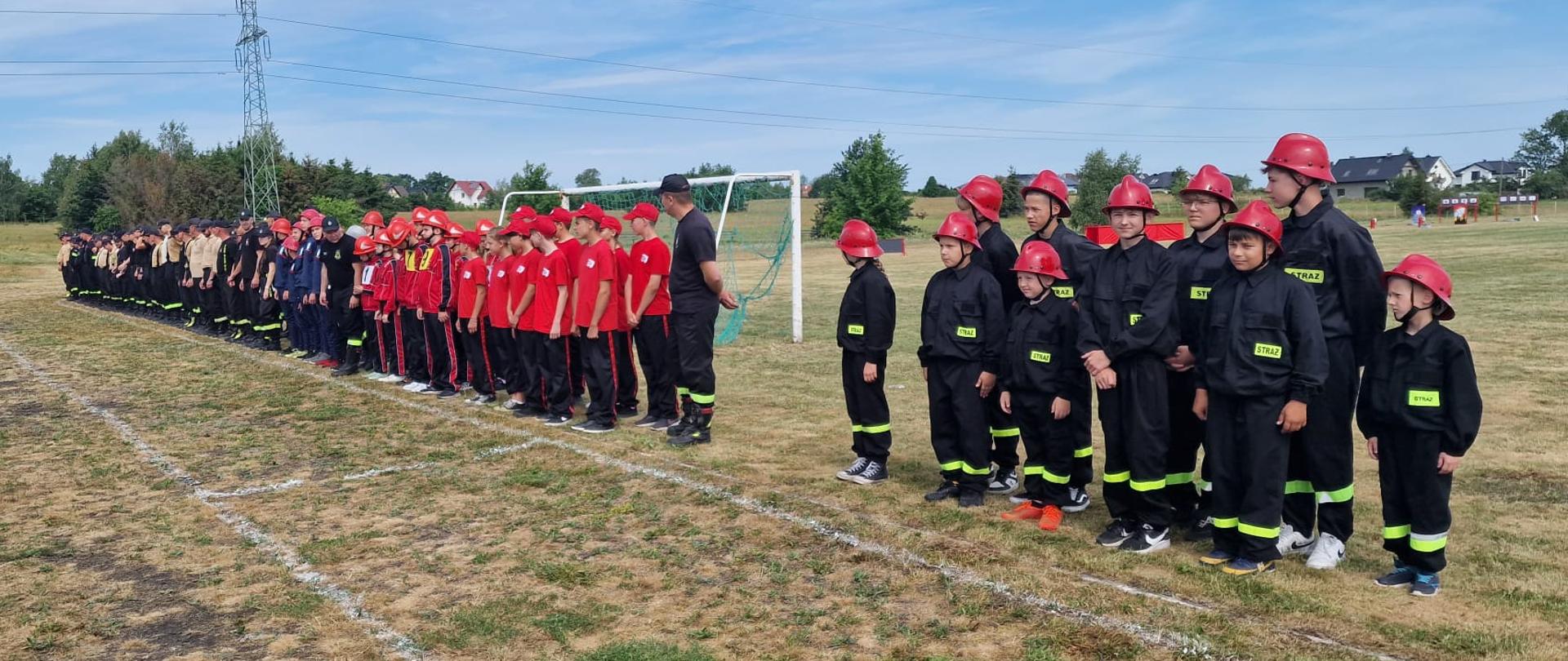 Zbiórka drużyn w dwuszeregu na boisku. Stoją zawodnicy MDP oraz OSP.