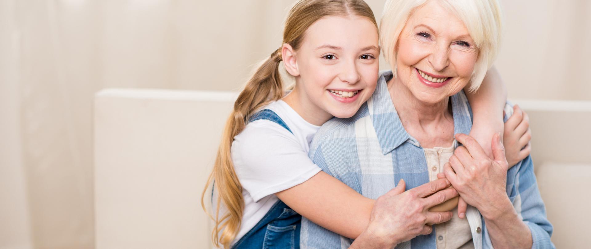 Na zdjęciu jest młoda dziewczyna, która obejmuje starszą kobietę. Siedzą na kanapie, są uśmiechnięte.