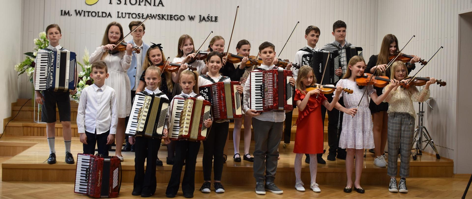 Na zdjęciu znajduje się grupa dzieci ze skrzypcami i akordeonami, stoją na scenie w auli PSM, w tle na jasnej ścianie logo szkoły oraz godło państwowe