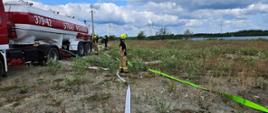 Ćwiczenia strażackie na otwartym terenie. W kilku miejscach widać prądy wody, strażaków przy pojazdach i zbiornikach oraz kilka wozów strażackich i cysternę. W tle rozległe pole i słupy energetyczne.