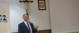 Minister Czarnek stoi za drewnianą mównicą i mówi do mikrofonu, za nim na ścianie godło i krzyż.