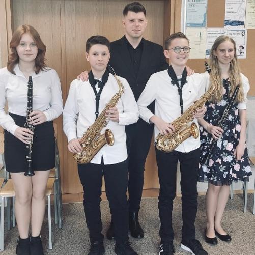 Na zdjęciu widzimy czworo uczniów, po środku stoi nauczyciel. Od lewej: Lena Kosielska (klarnet), Adrian Płochocki (saksofon), Mateusz Kurkowski (nauczyciel), Bartosz Surażyński (saksofon), Zuzanna Tarnacka (klarnet). Galowo ubrani uczniowie, trzymają w dłoniach instrumenty.