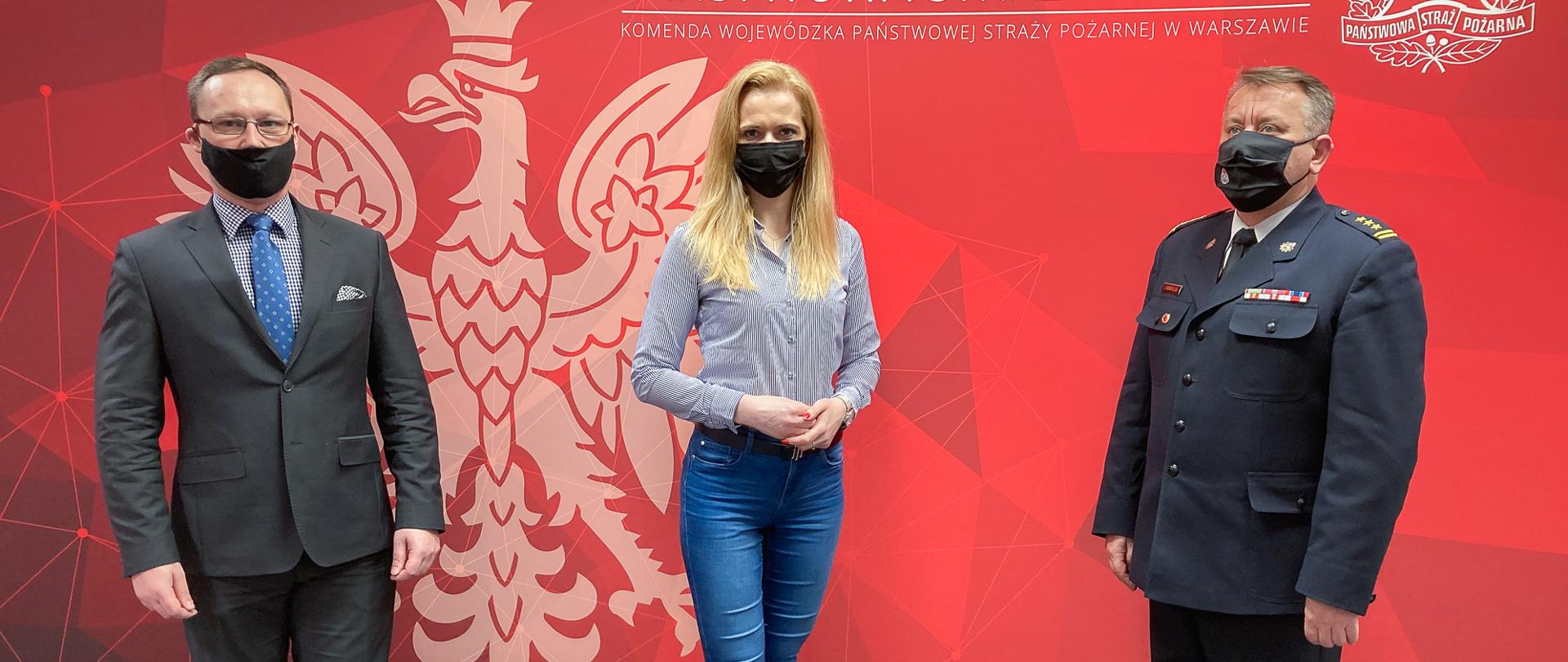 Zdjęcie przedstawia trzy osoby,w tym jednego strażaka, które stoją na tle czerwonej ścianki w dużym godłem RP koloru białego.