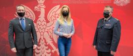 Zdjęcie przedstawia trzy osoby,w tym jednego strażaka, które stoją na tle czerwonej ścianki w dużym godłem RP koloru białego.