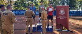 Zawodnicy stoją na podium medalowym z dyplomami w rękach i medalami na szyjach. Dwóch strażaków wręcza puchar. Obok nich stoi baner z logo PSP i napisem KOmenda Miejska PAństowej Straży Pożarnej w Rybniku. 