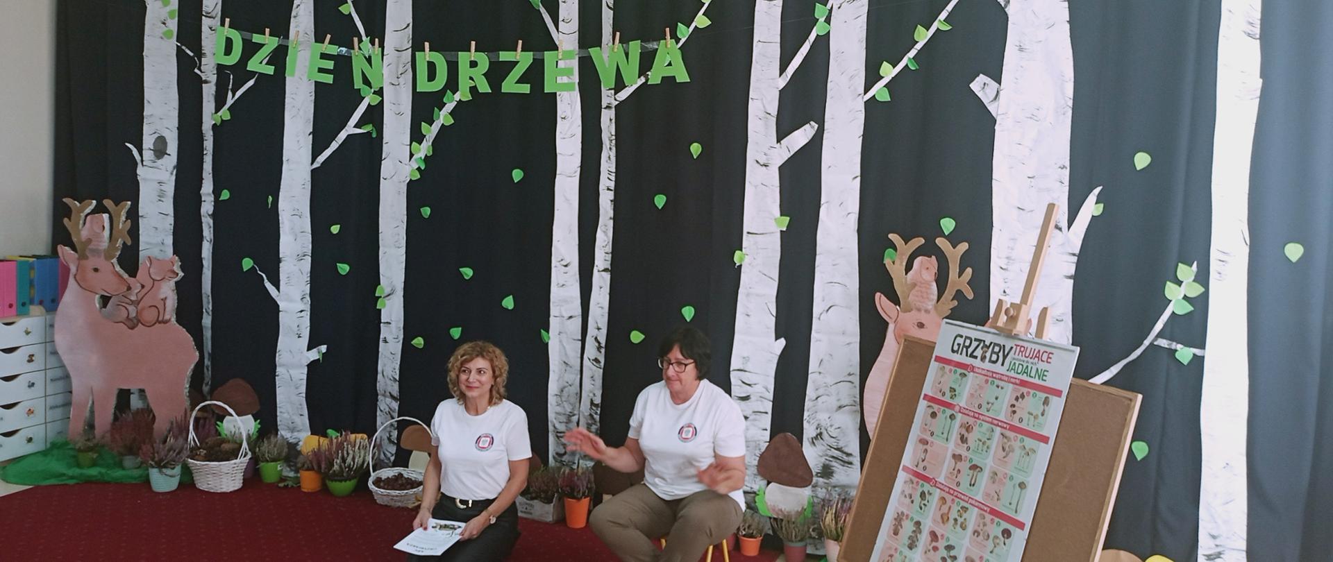 Dwóch pracowników PSSE w Garwolinie w czasie spotkania edukacyjnego w sali Przedszkola nr 6, w scenerii wykonanych różnymi technikami drzew, zwierząt, grzybów - w doniczkach wrzosy, obok plakat "Grzyby trujące i jadalne". Napis DZIEŃ DRZEWA