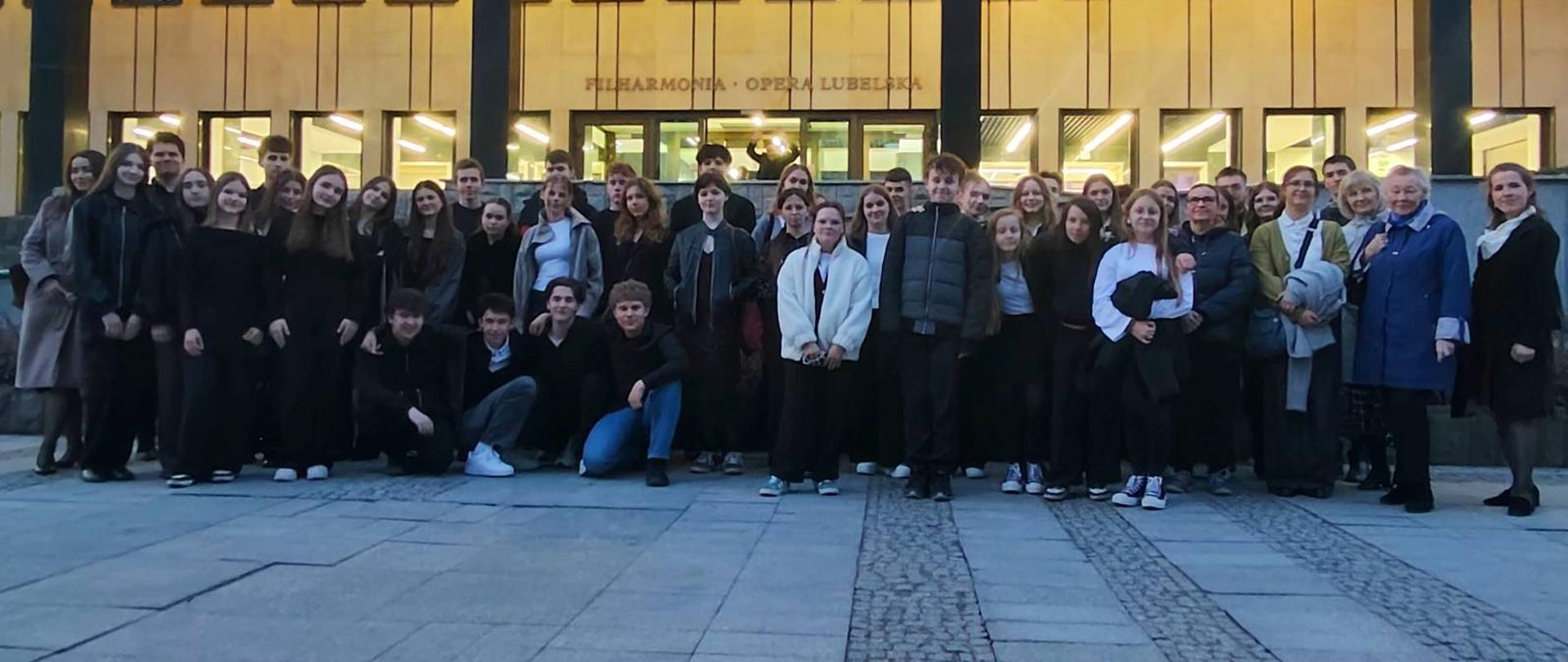 Grupa uczniów i nauczycieli stojąca przed budynkiem Filharmonii Lubelskiej podczas wyjścia na koncert „Pasji wg św. Jana” Jana Sebastiana Bacha.