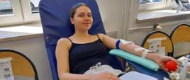 Na zajęciu widoczna kobieta w granatowej koszulce i białych spodniach siedząca na fotelu na sali oddający krew podczas akcji zbiórki krwi zorganizowanej przez Klub Honorowych Dawców Krwi Polskiego Czerwonego Krzyża przy Komendzie Powiatowej Państwowej Straży Pożarnej w Gorlicach i Klub Honorowych Dawców Krwi Polskiego Czerwonego Krzyża Moszczenica.