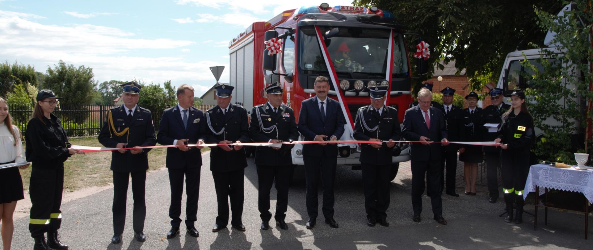 Zdjęcie przedstawia uroczystość przecięcia wstęgi przez ministra, posła, komendanta oraz druhów podczas przekazania nowego samochodu dla OSP Barłogi