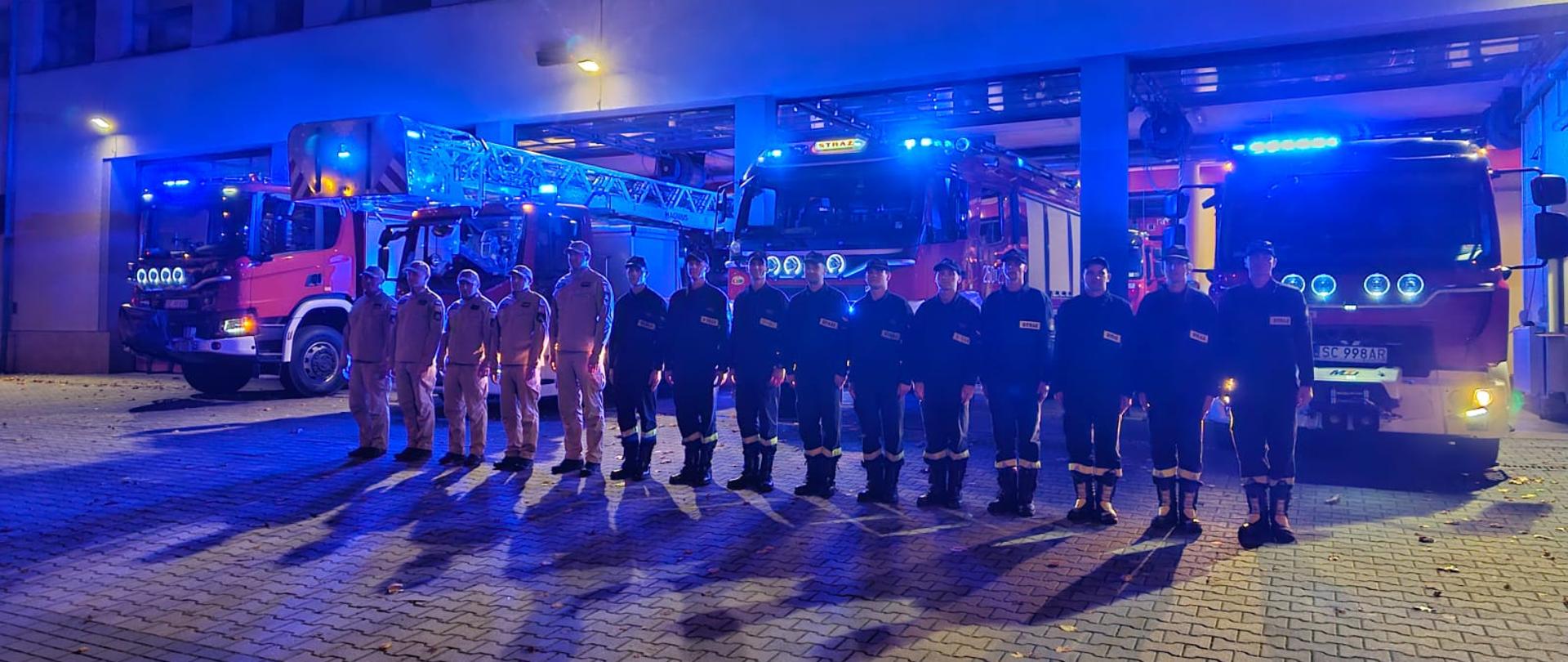 Strażacy ze zmiany JRG ustawieni w szeregu przed bramami garażowymi jednostki, za nimi widoczne samochody bojowe z włączonymi sygnałami świetlnymi