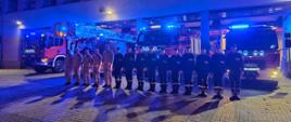 Strażacy ze zmiany JRG ustawieni w szeregu przed bramami garażowymi jednostki, za nimi widoczne samochody bojowe z włączonymi sygnałami świetlnymi