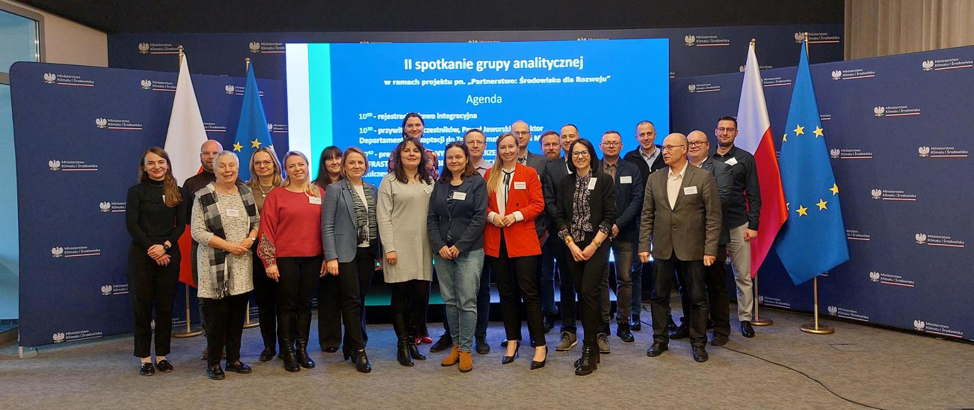 Zdjęcie zbiorowe przedstawicieli samorządów, instytucji i urzędów wykonane w sali konferencyjnej Ministerstwa Klimatu i Środowiska. Członkowie grupy stoją na tle telebimu z nazwą: II spotkanie grupy analitycznej w ramach projektu pn. "Partnerstwo: Środowisko dla Rozwoju".