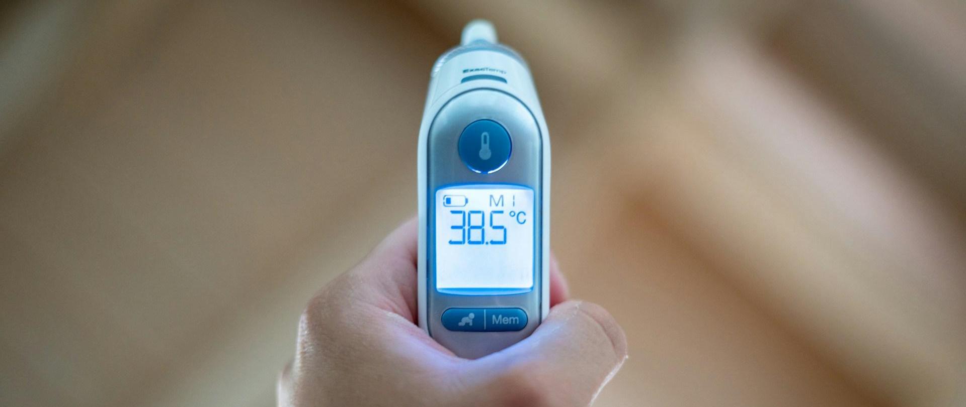 Dłoń trzymająca elektroniczny termometr ze wskazaniem 38,5 stopnia Celsjusza.