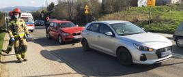 Dwa samochody osobowe uczestniczące w zdarzeniu stoją jeden za drugim na jednym pasie drogi. Jedn samochód koloru jasnego a drugi czerwonego Obok stoją strażacy. W tle karetka Pogotowia Ratunkowego.