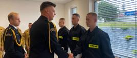 W sali szkoleniowej jednostki ratowniczo- gaśniczej Komendant Miejski ściska rękę nowozatrudnionemu funkcjonariuszowi. Dookoła znajdują się inni funkcjonariusze w mundurach. 