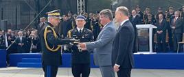 Sekretarz stanu w Ministerstwie Spraw Wewnętrznych i Administracji wręcza szablę pomorskiemu komendantowi Państwowej Straży Pożarnej. Obok stoi prezydent Rzeczpospolitej Polskiej, z drugiej strony strażak. Wszyscy stoją przed trybuną honorową na której są zaproszeni goście.