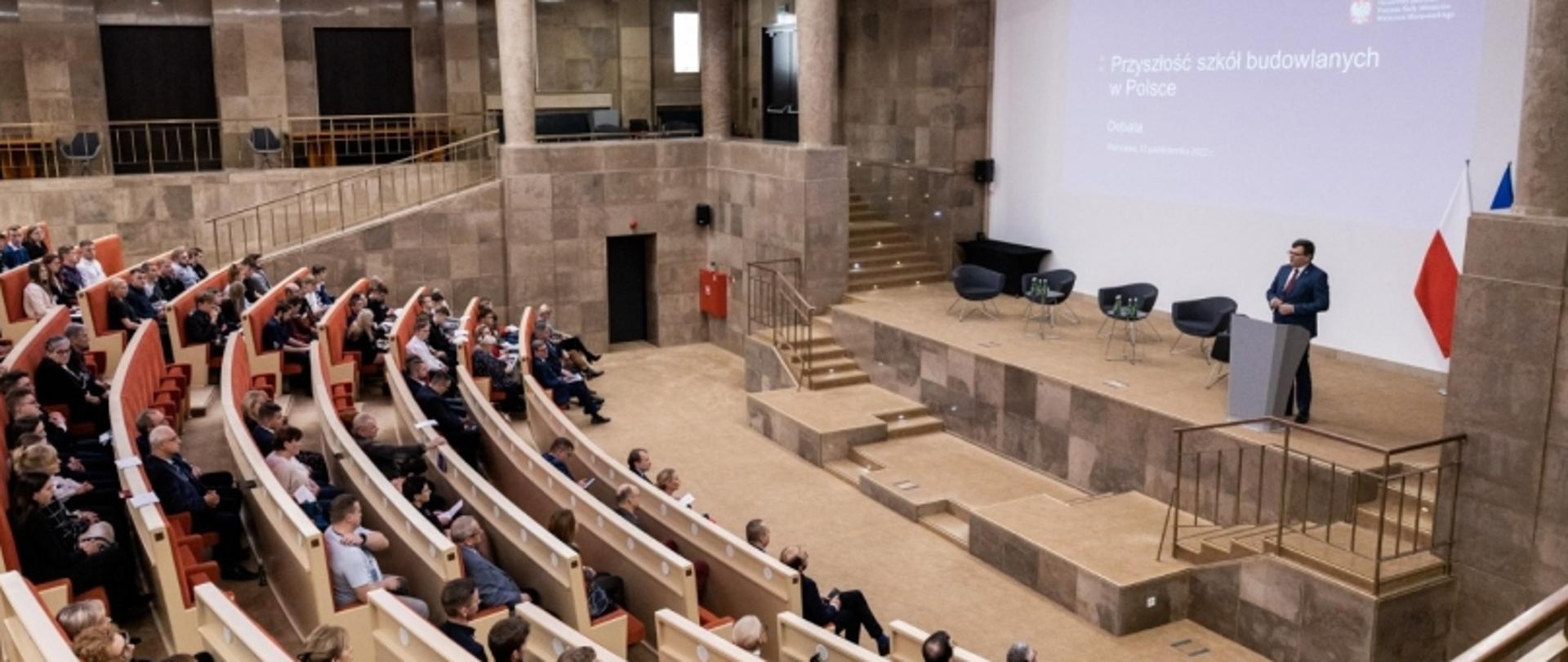 Prelegent na scenie w auli konferencyjnej, za nim ekran z napisem „Przyszłość szkół budowlanych w Polsce” oraz flagi Polski i UE, widownia siedzi w rzędach.