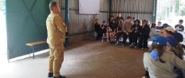 Zdjęcie przedstawia uczestników obozu harcerskiego pod namiotami oraz kadrę opiekunów podczas pogadanki edukacyjnej dla dzieci, młodzieży i osób dorosłych prowadzonej przez funkcjonariusza Komendy Powiatowej Państwowej Straży Pożarnej w Gołdapi