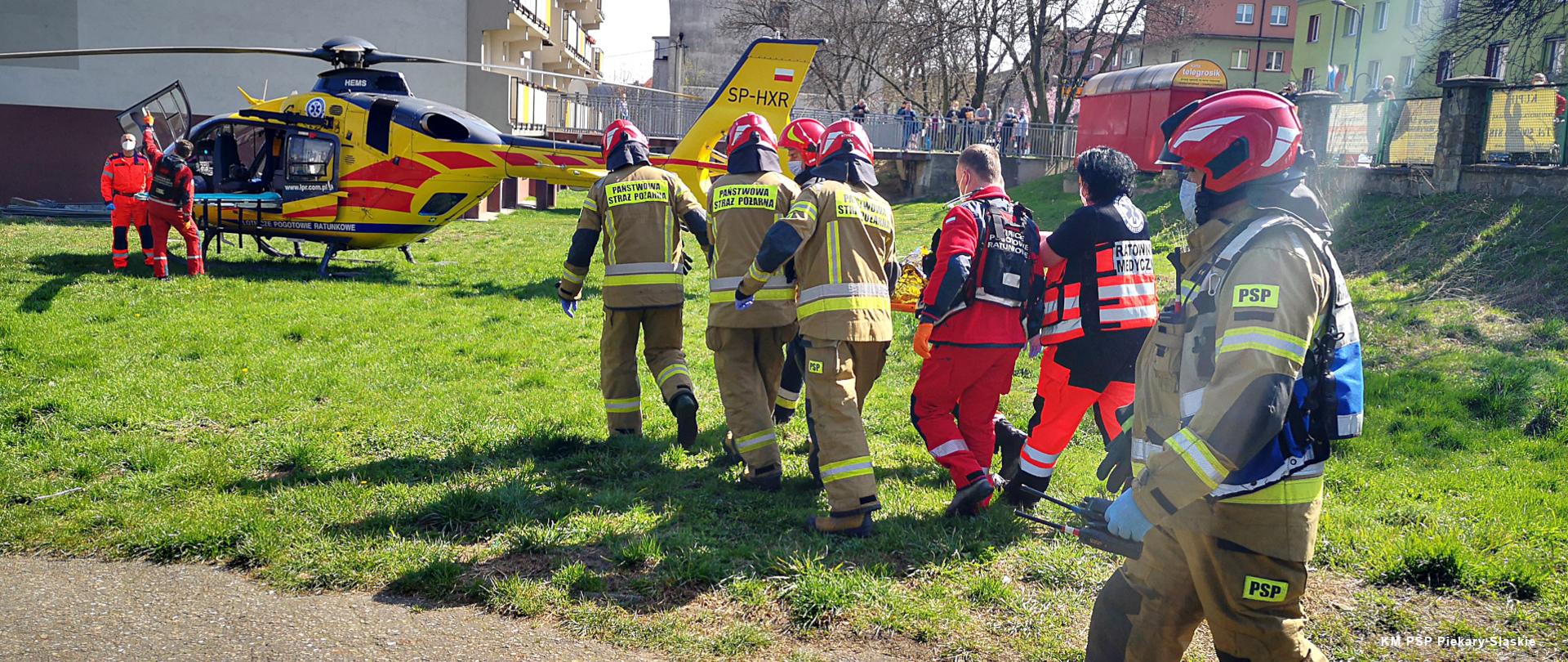 Zdjęcie przedstawia na pierwszym planie 5 strażaków PSP oraz 3 ratowników PRM transportujących poszkodowanego do stojącego nieopodal śmigłowca LPR obok którego stoją pilot i ratownik medyczny.