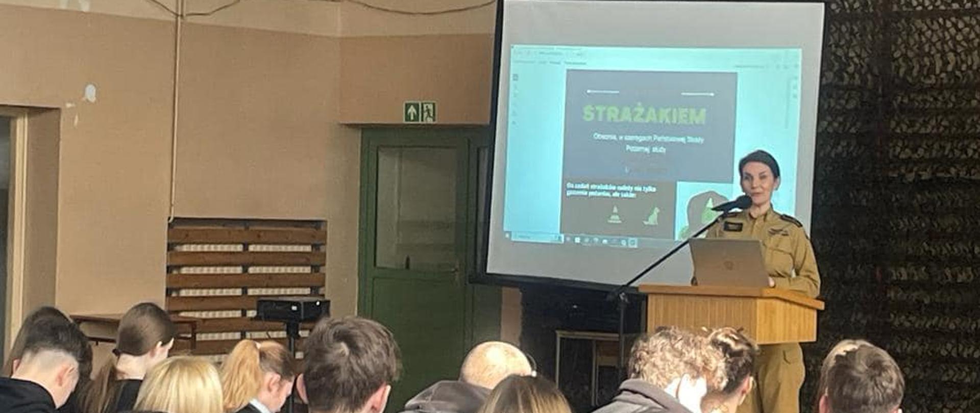 Na zdjęciu znajduje się funkcjonariuszka straży pożarnej stojąca za mównicą, w tle widać slajd na ekranie, na sali znajdują się osoby ubrane w mundury
