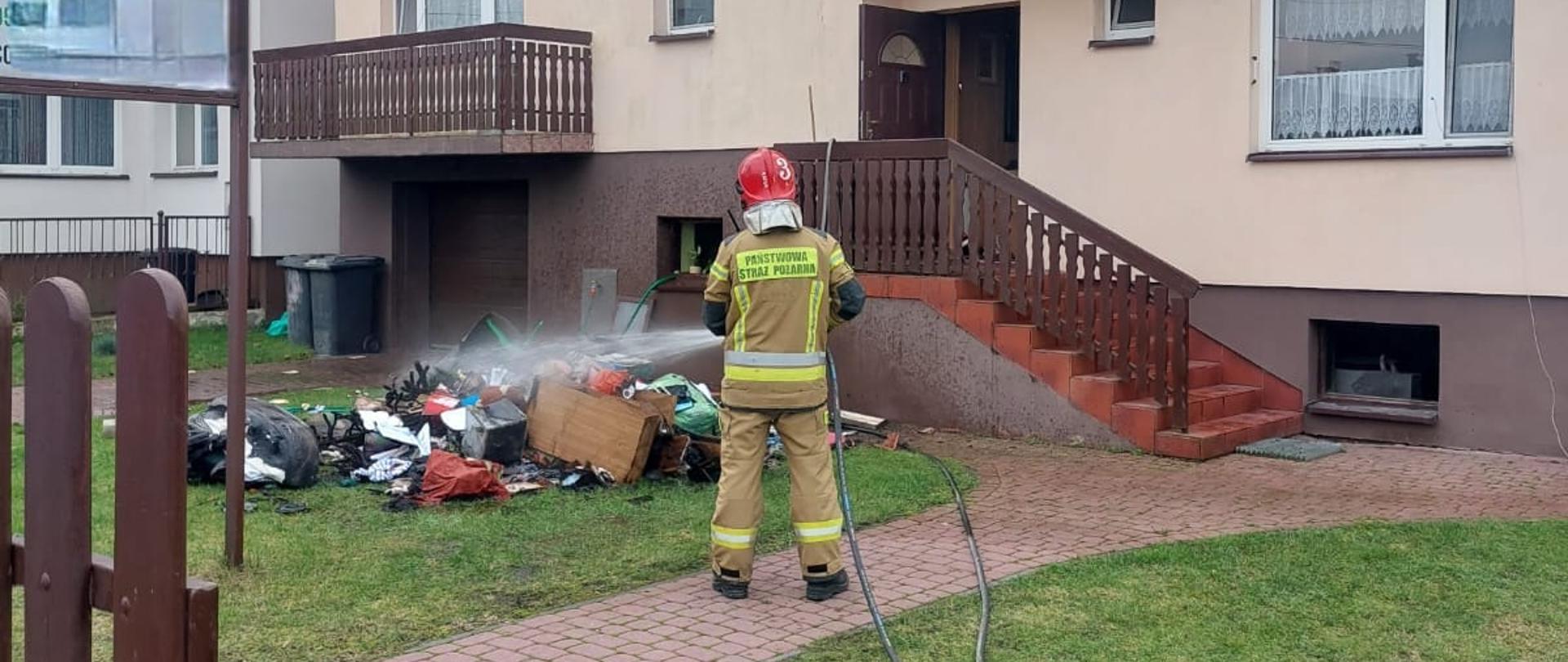 Strażak w umundurowaniu specjalnym na tle budynku jednorodzinnego gaszący nadpalone elementy wyposażenia na zewnątrz budynku.