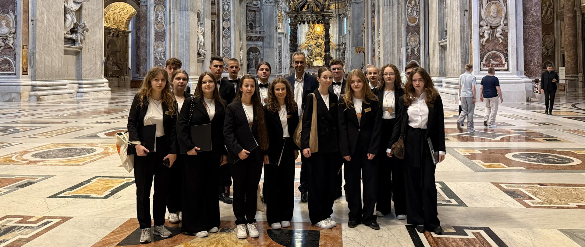 Członkowie chóru w czarnych garniturach i z teczkami na nuty stoją wewnątrz bogato zdobionej Bazyliki św. Piotra w Watykanie. W tle widać monumentalne kolumny, rzeźby oraz wznoszącą się w głębi kopułę.