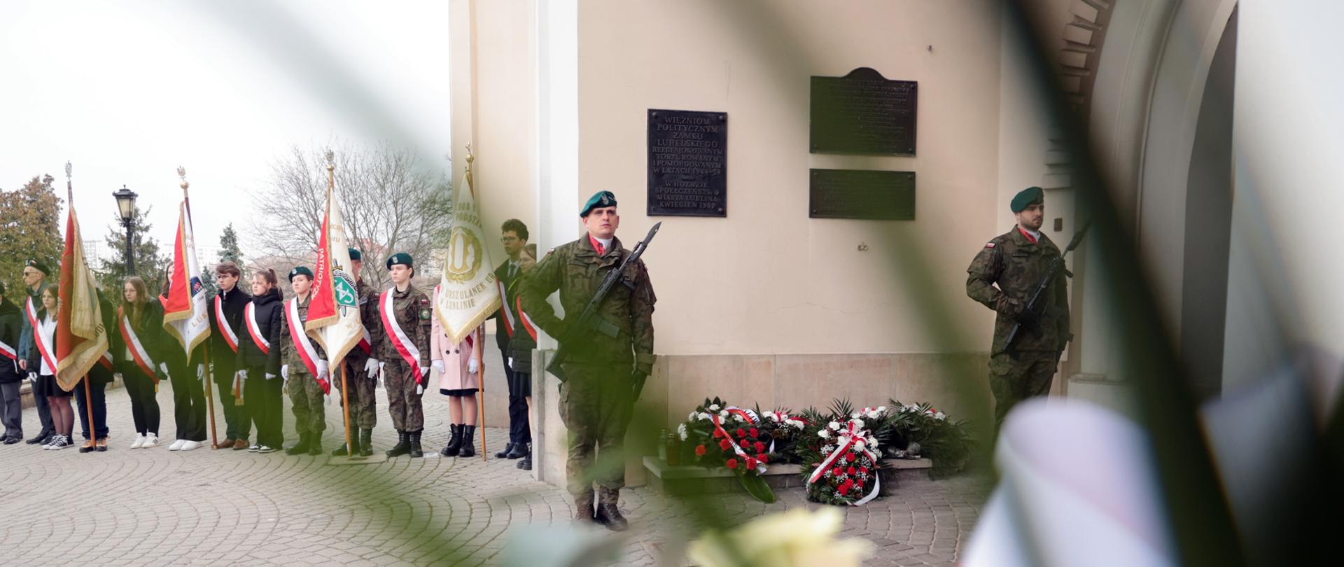Dwóch żołnierzy stoi przy tablicy upamiętniającej. Pod tablicą kwiaty.