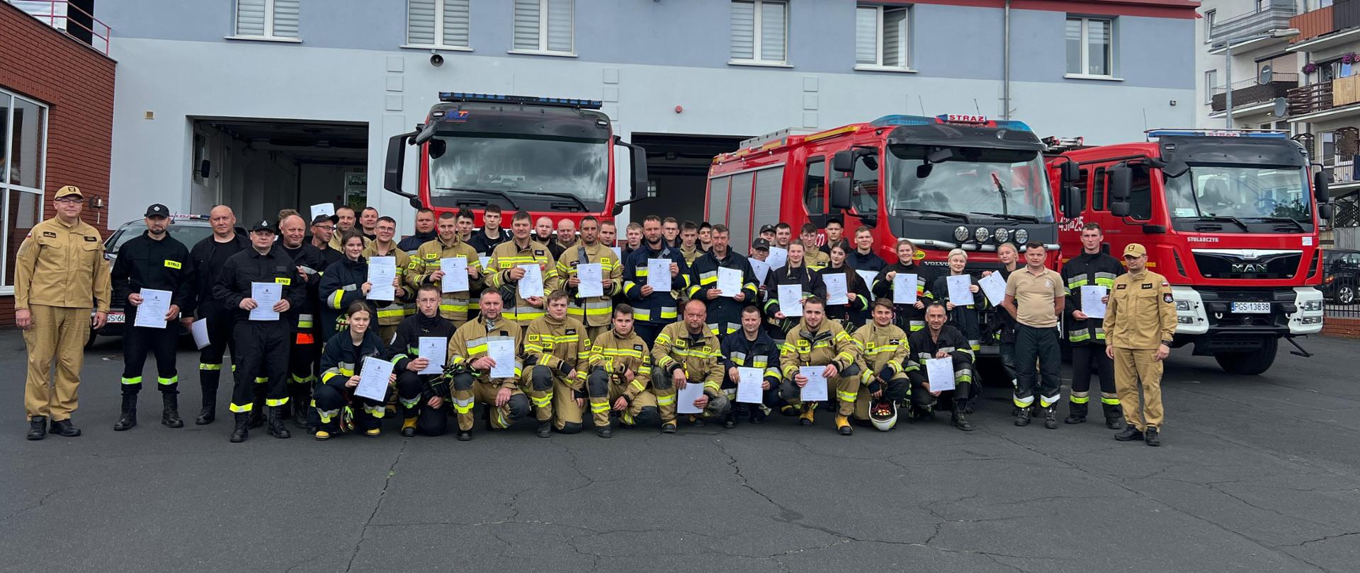 Wspólne zdjęcie wszystkich kursantów wraz z komendantem przed komendą. W tle stoją pojazdy pożarnicze.