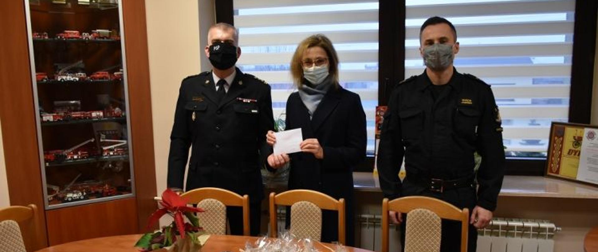 Na pierwszym planie od lewej komendant powiatowy PSP w Skarżysku-Kamiennej, opiekunka chorej dziewczynki trzymająca kopertę ze środkami finansowymi przekazanymi przez strażaków po prawej stronie zastępca komendanta powiatowego PSP. Na zdjęciu przed osobami znajduje się stolik na którym znajdują się pierniki przekazane przez opiekunkę dziewczynki w dowód wdzięczności za okazaną pomoc. Na stoliku stoi również kwiatek gwiazda betlejemska. W tle okno gabinetu oraz gablota.