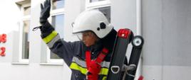 Widok z boku. Druhna OSP stojąca na drabinie pożarniczej z podniesioną ręką w trakcie ćwiczeń ze sprzętem.
