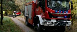 Samochód pożarniczy marki Iveco stoi na drodze gruntowej w lesie. W tyle zdjęcia trzech strażaków i przód samochodu Jelcz.