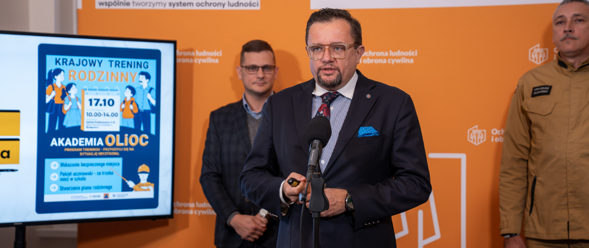 Irek Nitkiewicz przemawia do mikrofonu trzymanego na stojaku. W tle za nim stoi strażak w piaskowym mundurze z napisem „STRAŻ” na piersi. Obok znajduje się rolowana bannerowa ścianka z napisem „BEZPIECZNE KUJAWSKO-POMORSKIE”. 